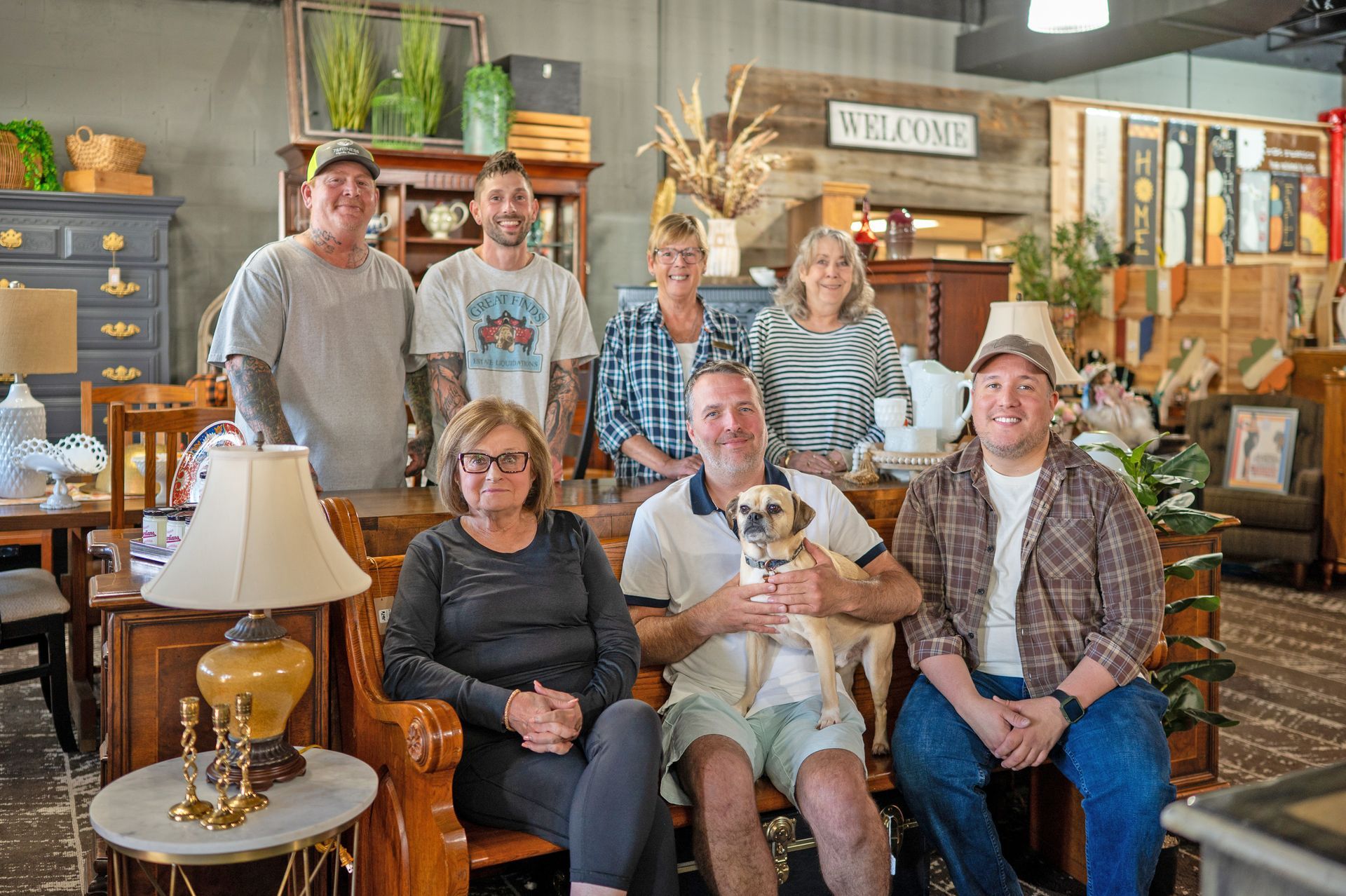 Group of seven people with a small dog posing in a home decor store, standing and sitting among furniture and accents.