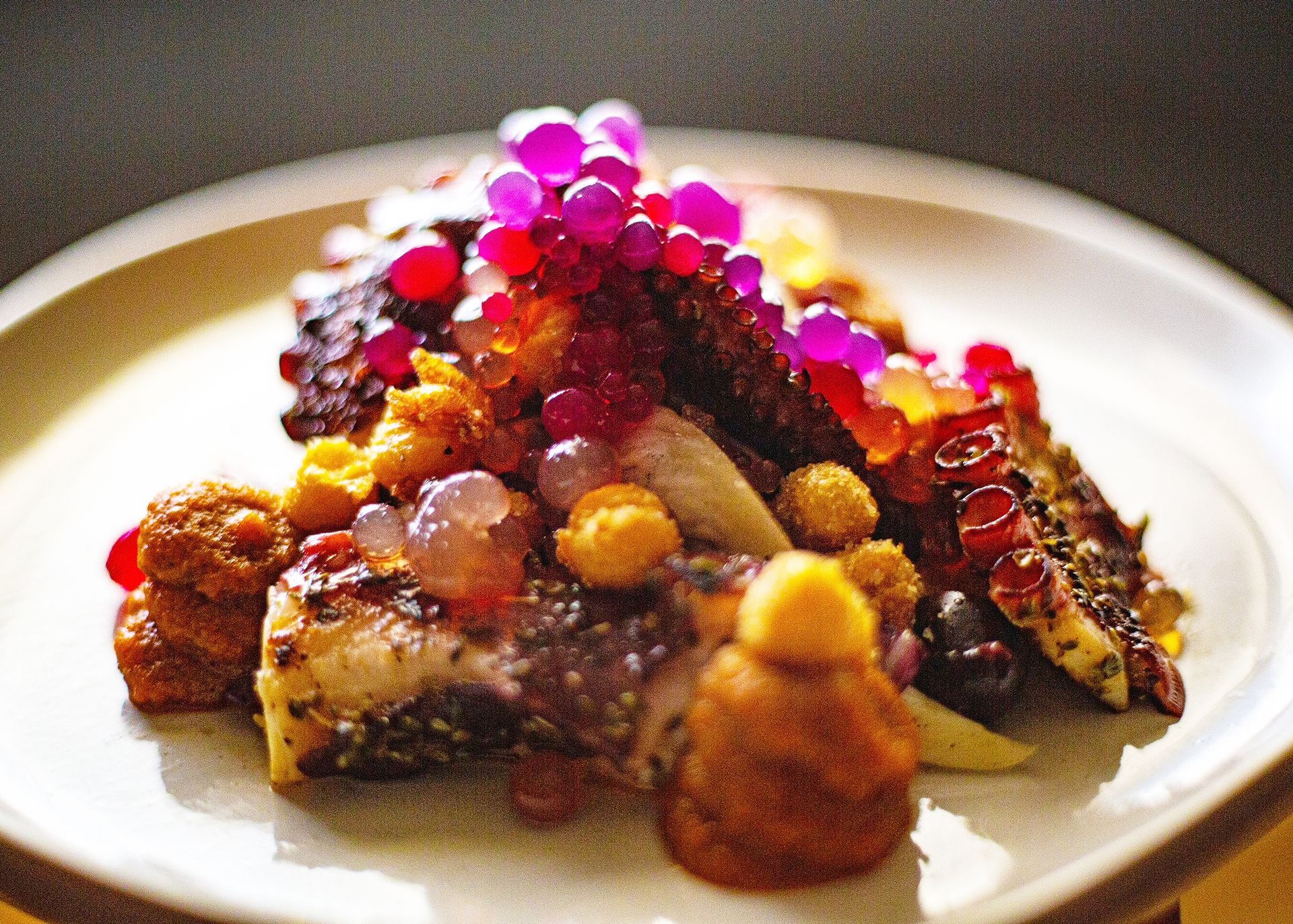 A plated dish featuring grilled octopus topped with vibrant pink and translucent pearls, served with small golden garnishes.