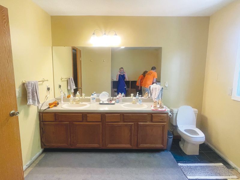 A bathroom with a double-sink wooden vanity, a large mirror reflecting two people, and a white toilet on the right.