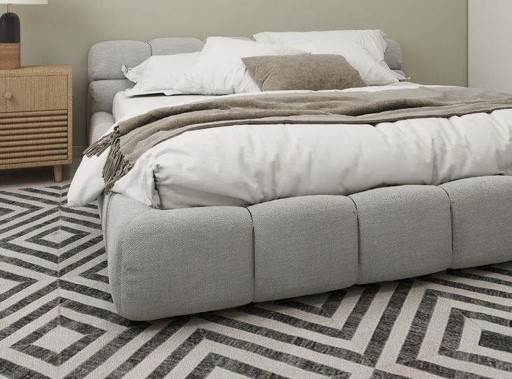 A plush, light gray upholstered bed with a modern, tufted frame sits on a patterned geometric rug beside a wood nightstand.