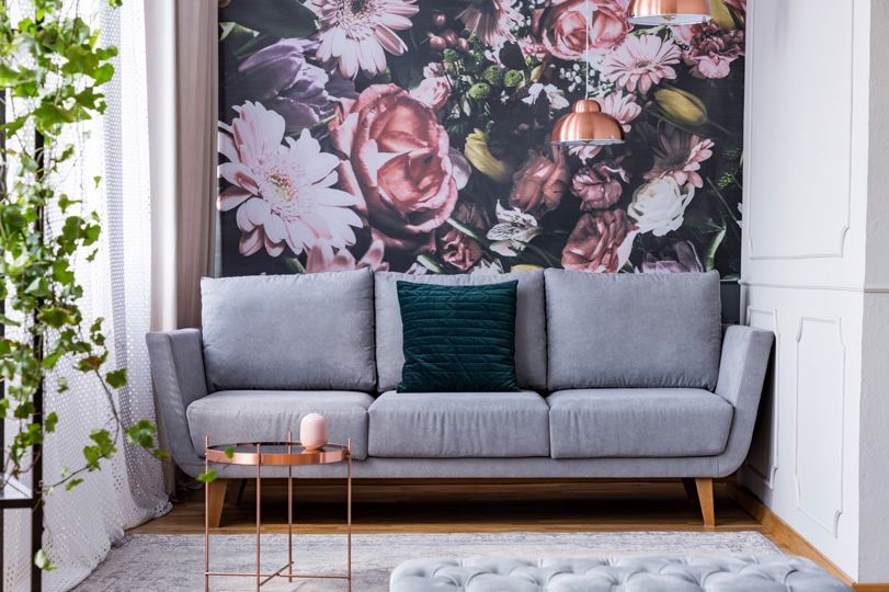 A grey sofa with a dark green pillow sits in front of a floral wallpaper accent wall next to a copper side table.