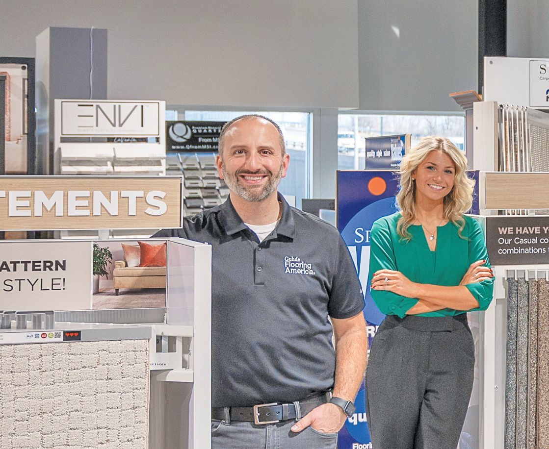 A man and a woman pose in a showroom featuring flooring and design samples, with shelves and marketing signage visible.