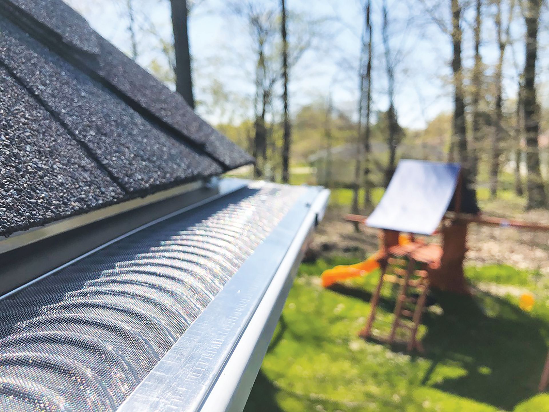 Close-up of a metal gutter guard installed on a roof, with a blurred backyard playground in the background.