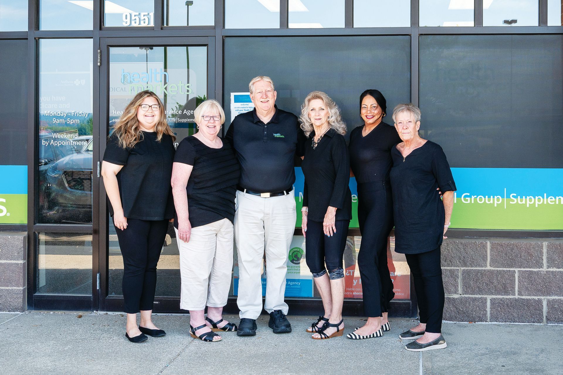 Six people stand in a line smiling in front of a storefront window with blue and green signage.