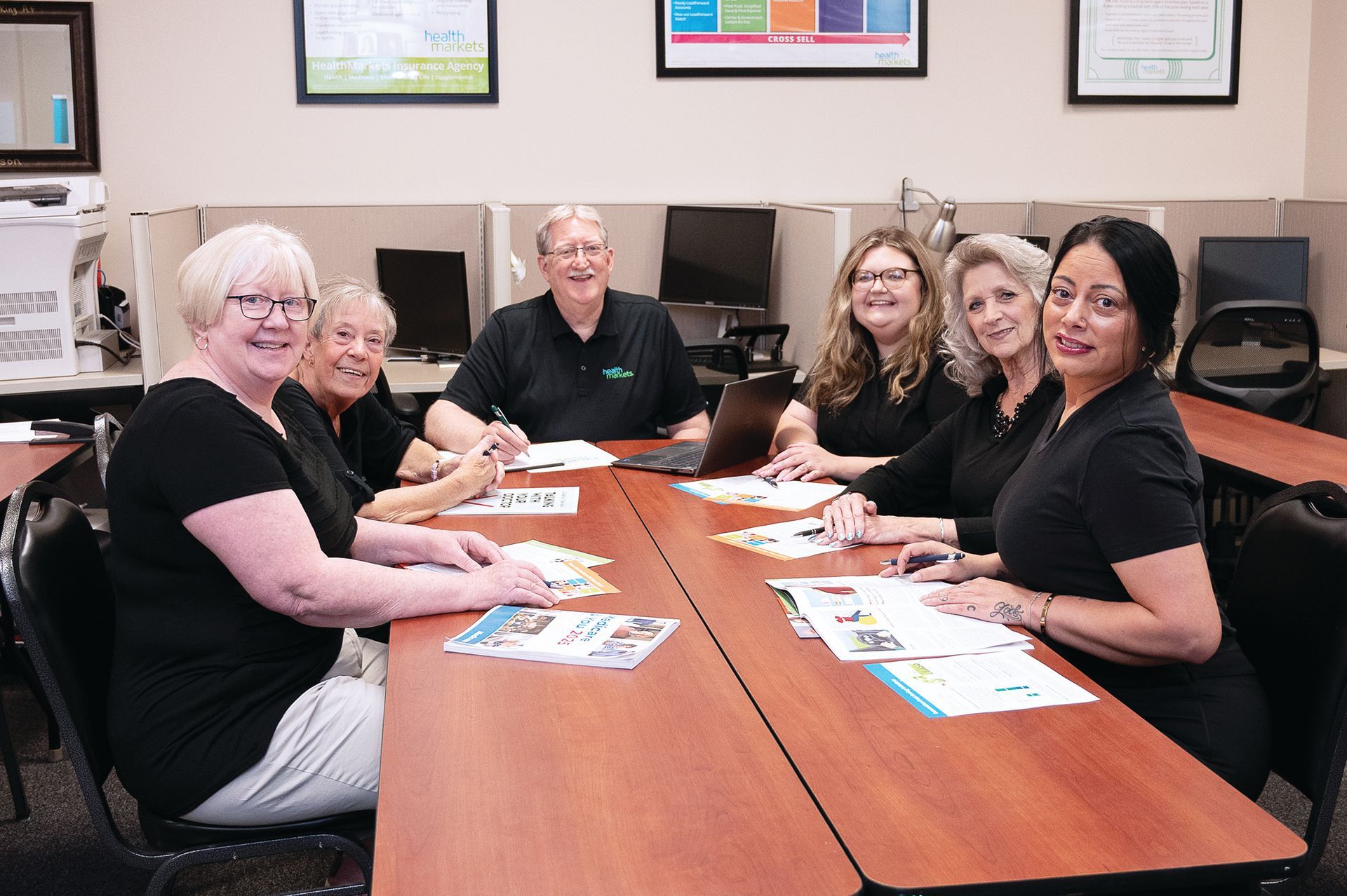 Six people sit around a conference table in an office, smiling at the camera with documents spread out before them.