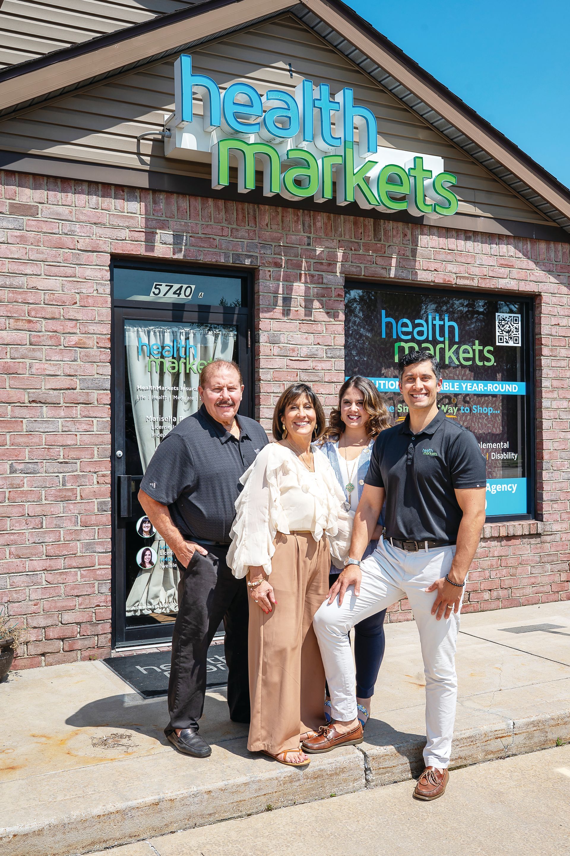 Four people stand together smiling outside a HealthMarkets office building with brick siding and glass entry doors.