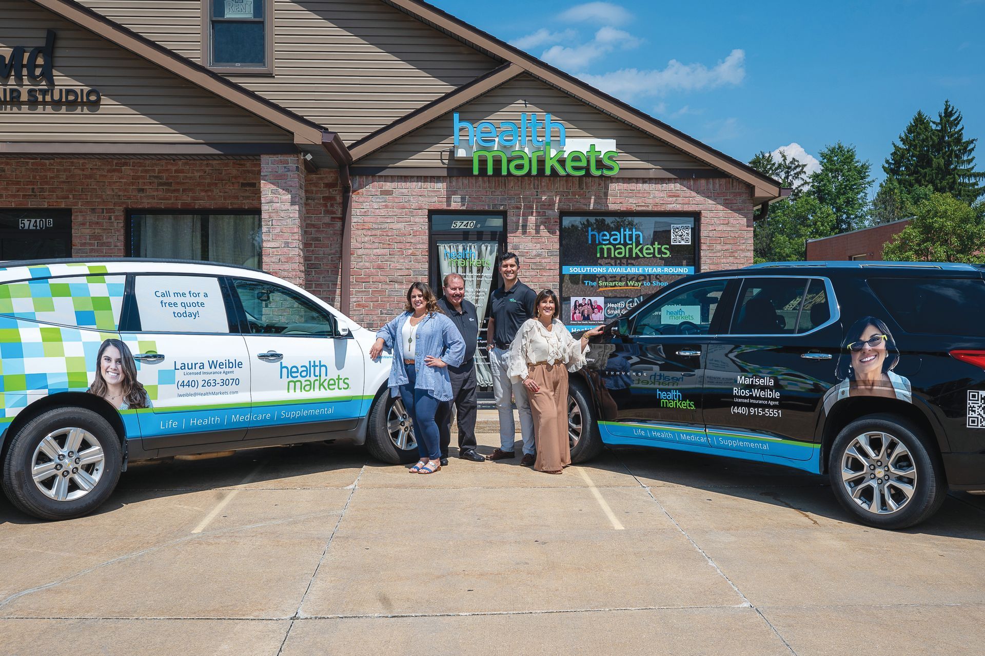 Four people pose outside a HealthMarkets office building between two branded company vehicles on a sunny day.