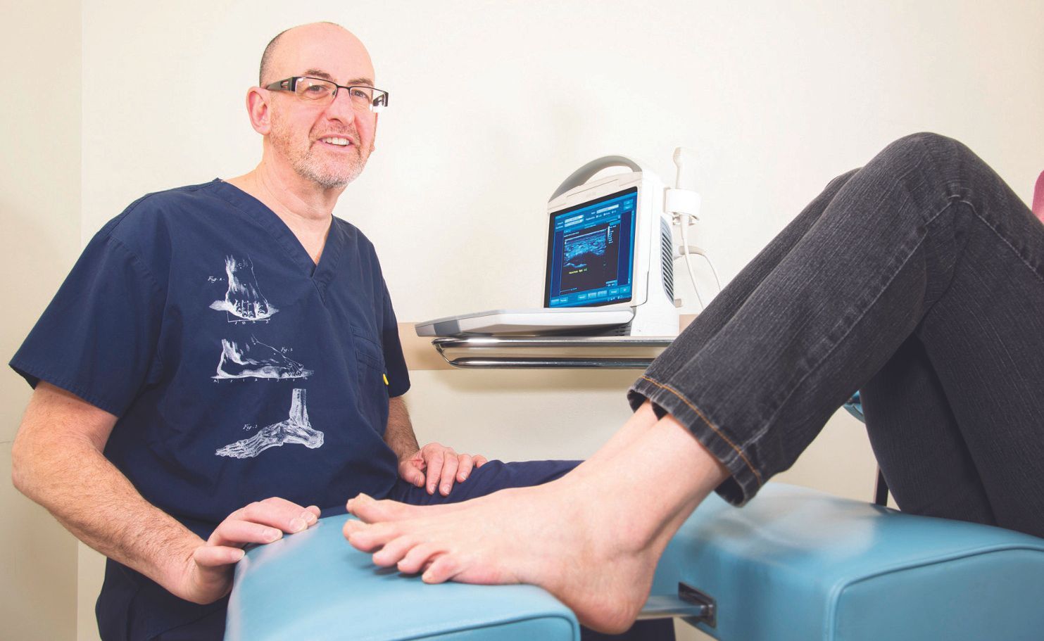 A healthcare provider in blue scrubs examines a patient's foot on a leg rest next to a portable ultrasound machine.