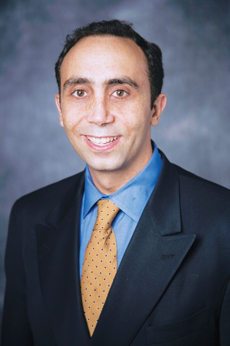 A headshot of a person wearing a dark blue suit, light blue shirt, and gold-dotted tie against a grey backdrop.