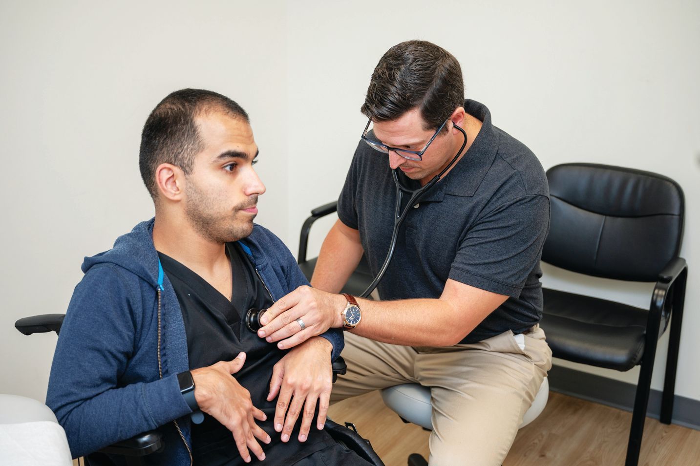 A healthcare professional using a stethoscope to examine a patient seated in a wheelchair in a medical office.