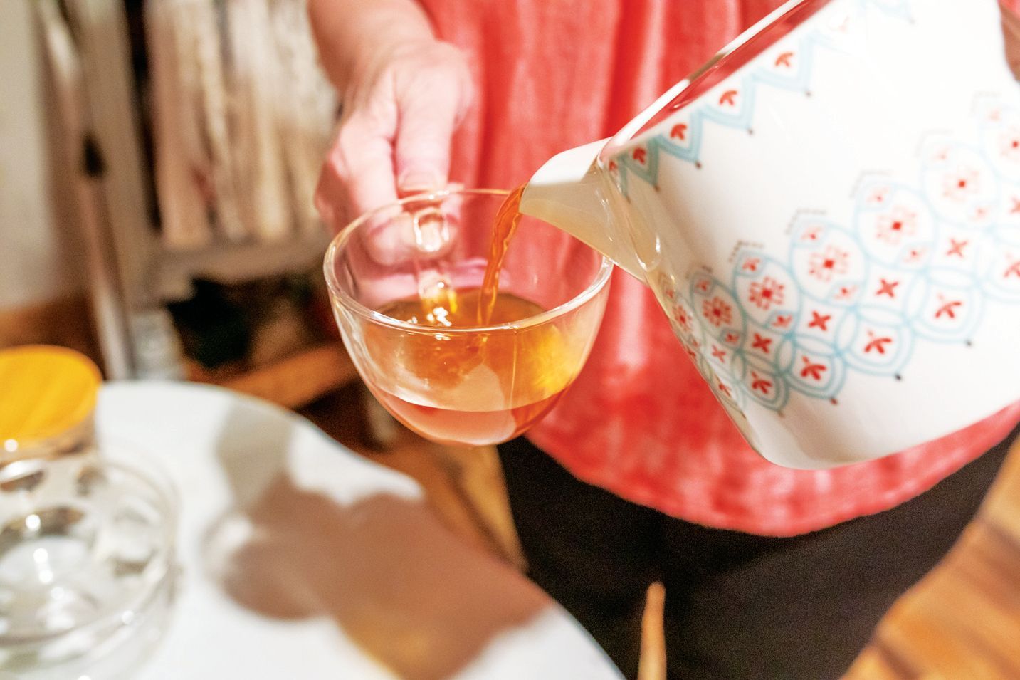 A person in a coral top pours tea from a patterned white pitcher into a clear glass cup.