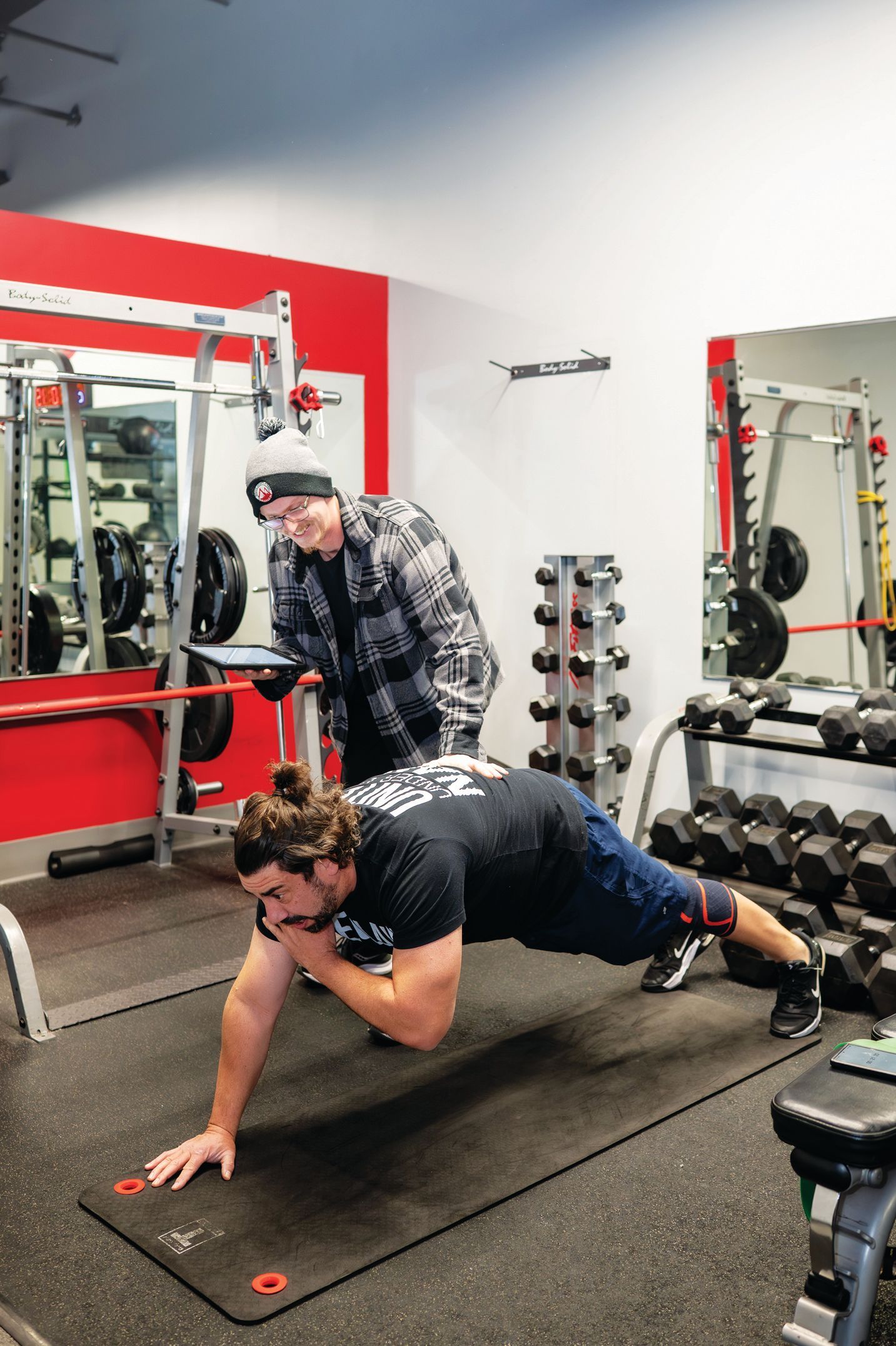 A person performs a plank in a gym while another person watches and holds a tablet.