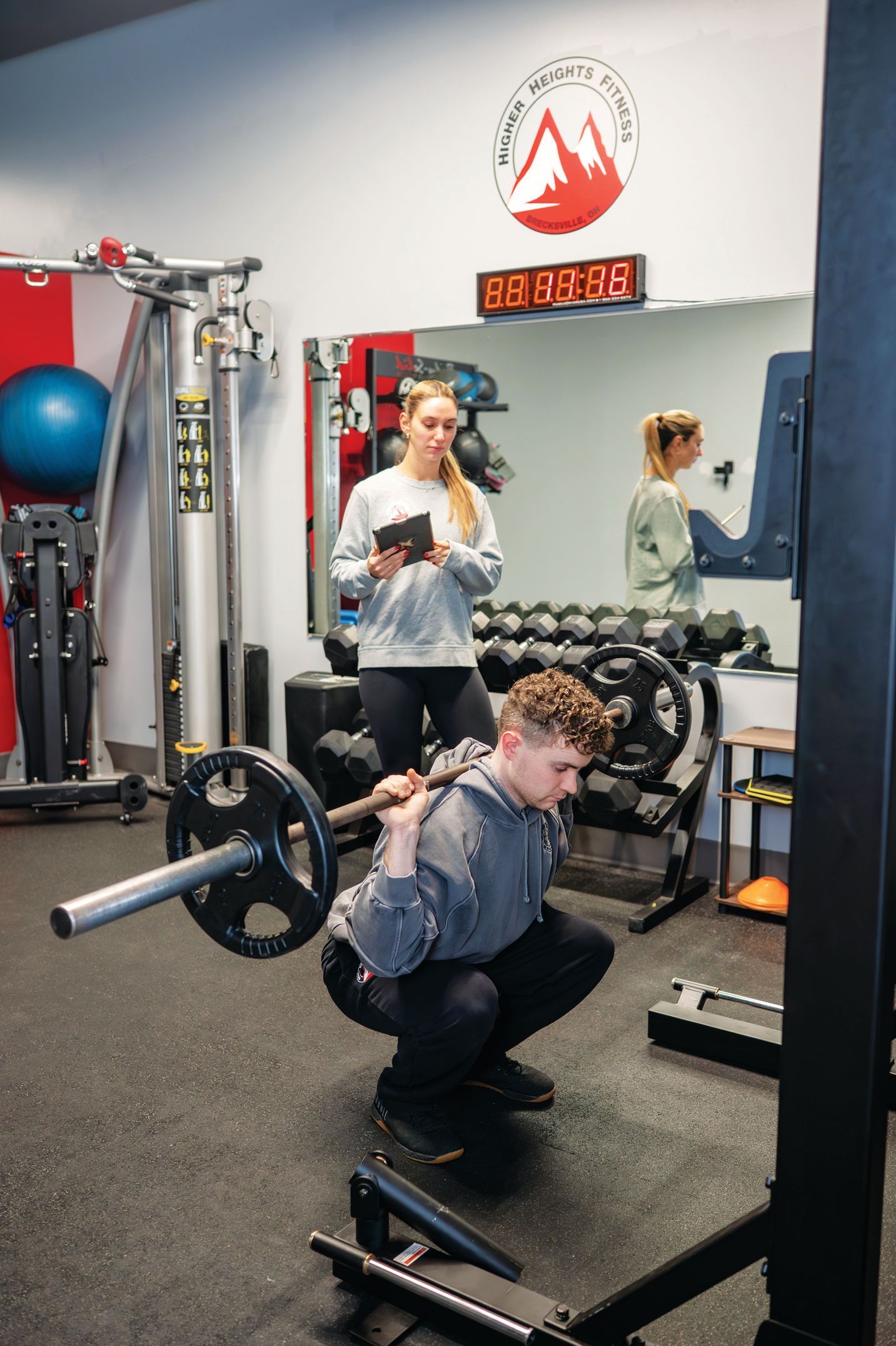 A person performs a barbell squat in a gym, observed by a trainer standing behind them.