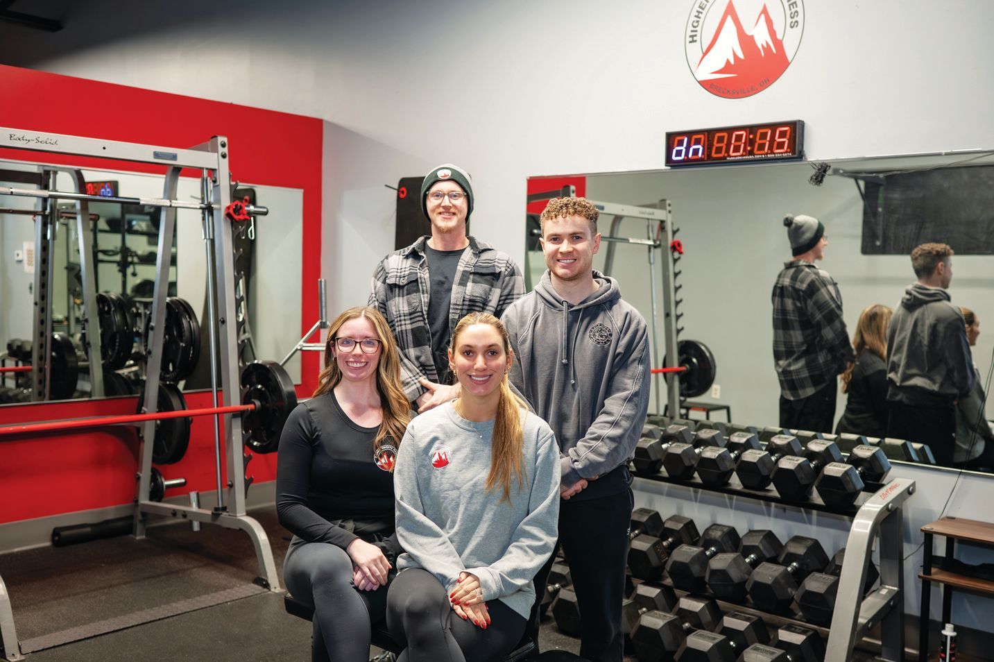 Four people posing for a group photo inside a gym with red walls, exercise equipment, and a mirror in the background.