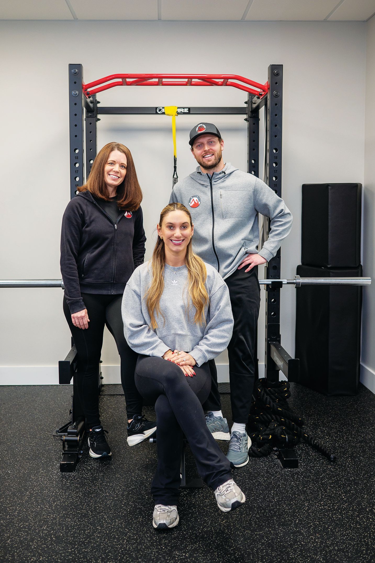 Three people stand and sit in front of a black gym power rack in an indoor space with light-gray walls and black flooring.