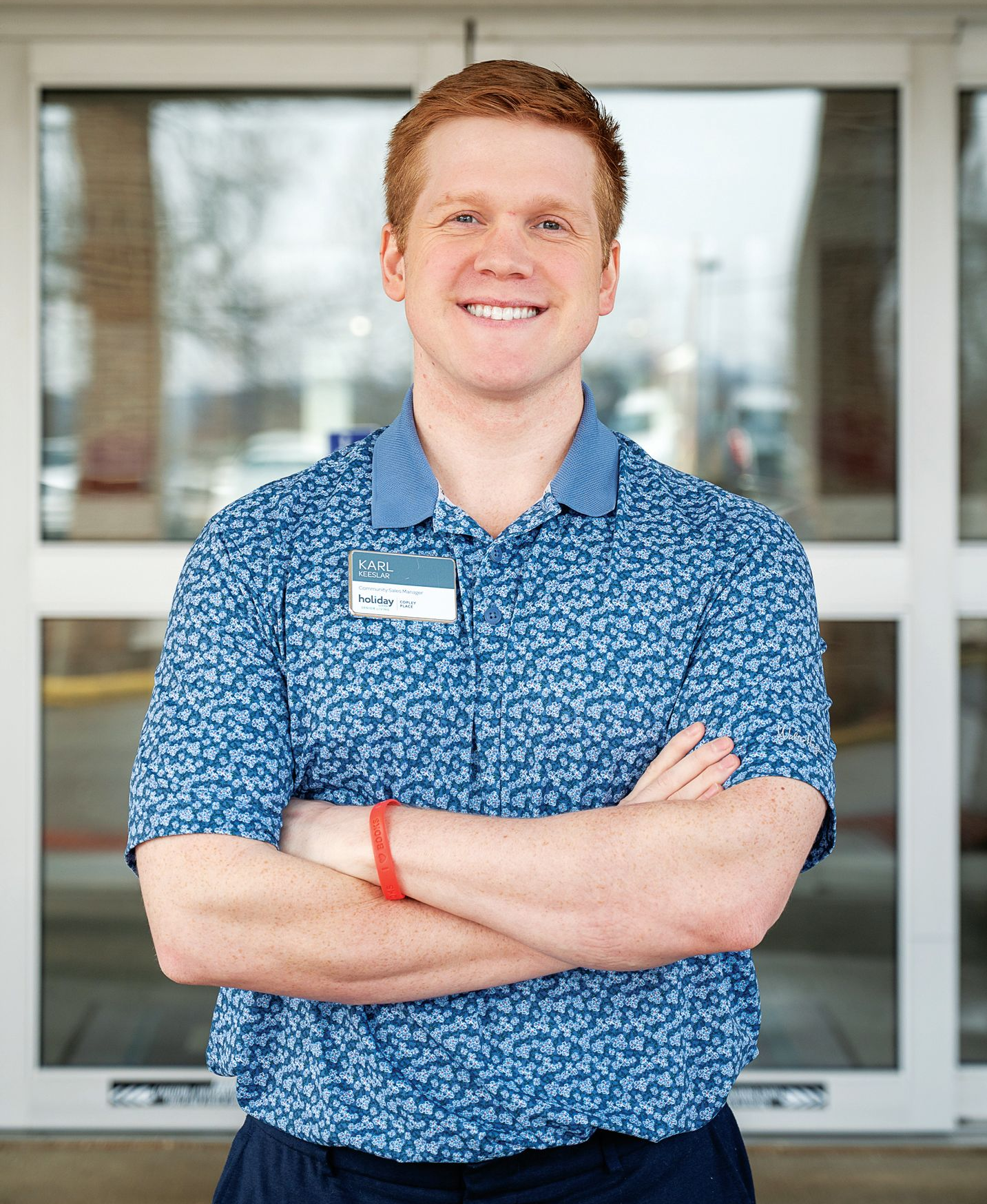 A smiling person with red hair, wearing a patterned blue polo shirt with a name tag, stands with arms crossed outdoors.