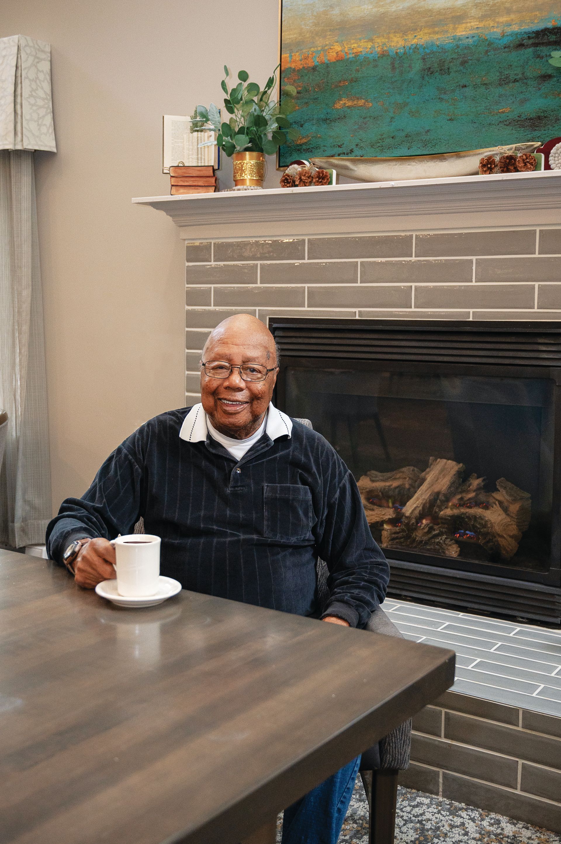 An individual sits at a wooden table holding a white coffee cup, with a brick fireplace and decor in the background.