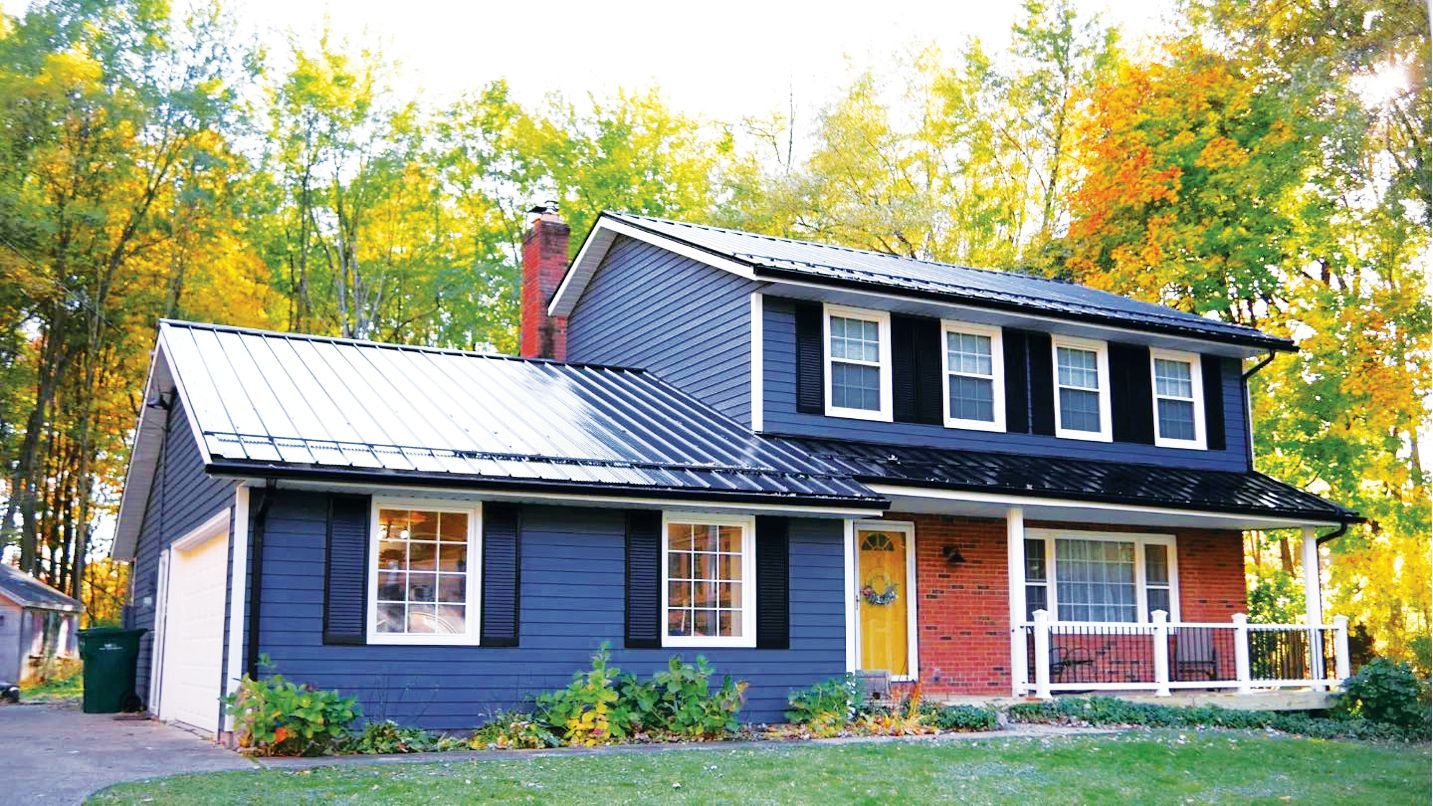 A two-story blue house with a dark metal roof, yellow front door, and trees with autumn foliage in the background.