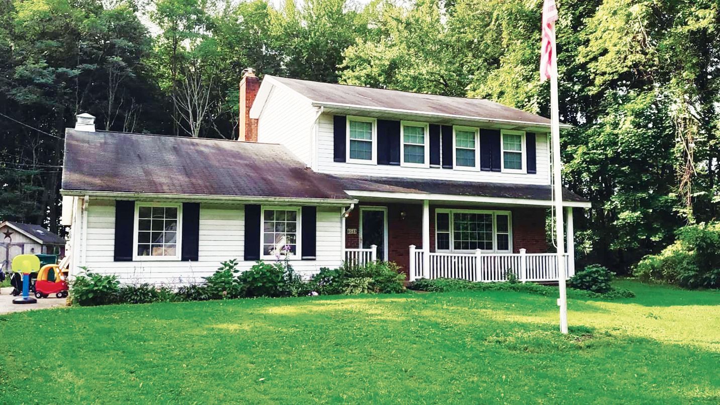 A two-story house with white siding and a brick lower level, surrounded by trees and a green lawn, with a tall flagpole.