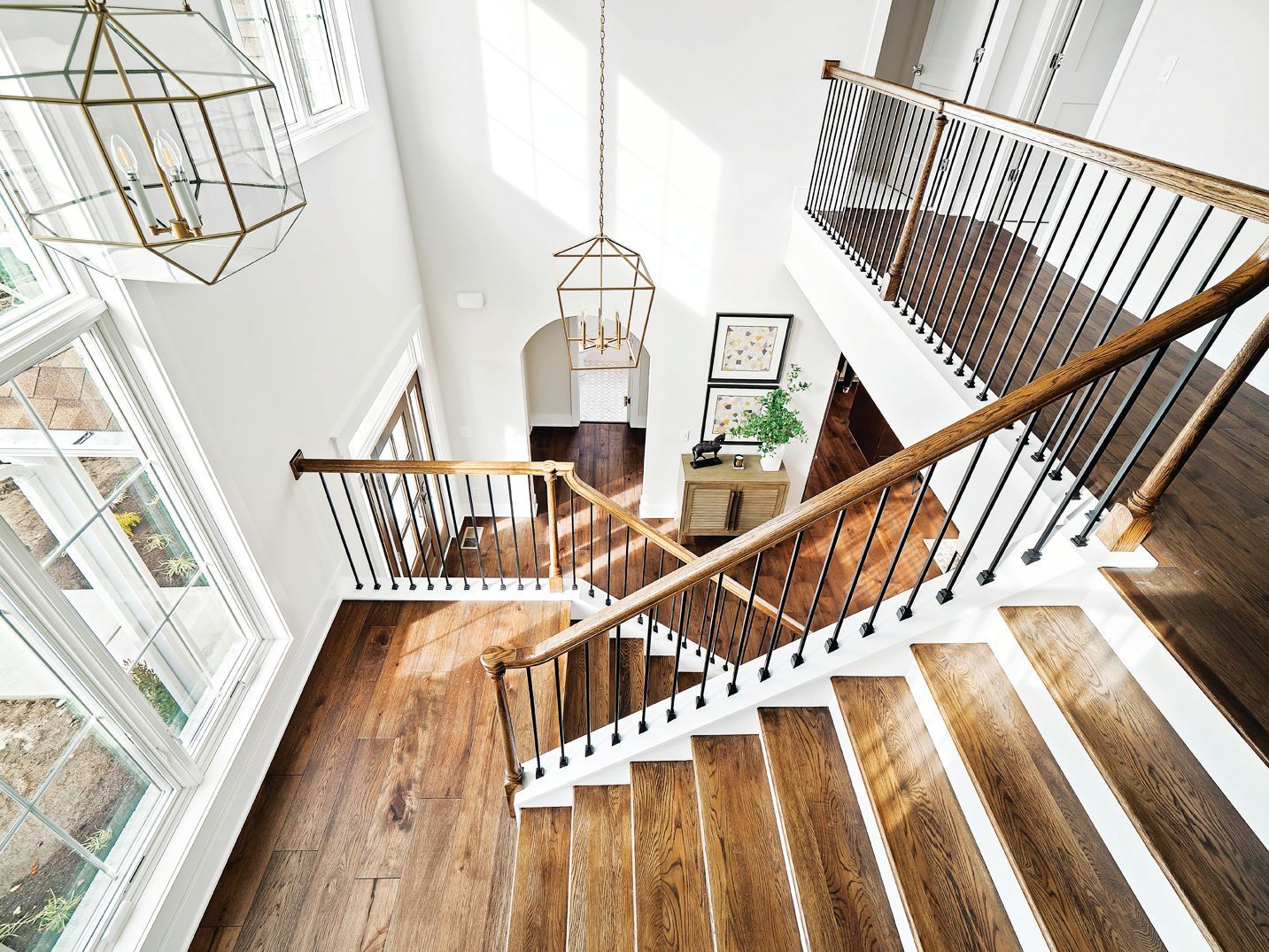 A high-angle view of a bright entryway with a wooden staircase, dark hardwood floors, white walls, and geometric lanterns.