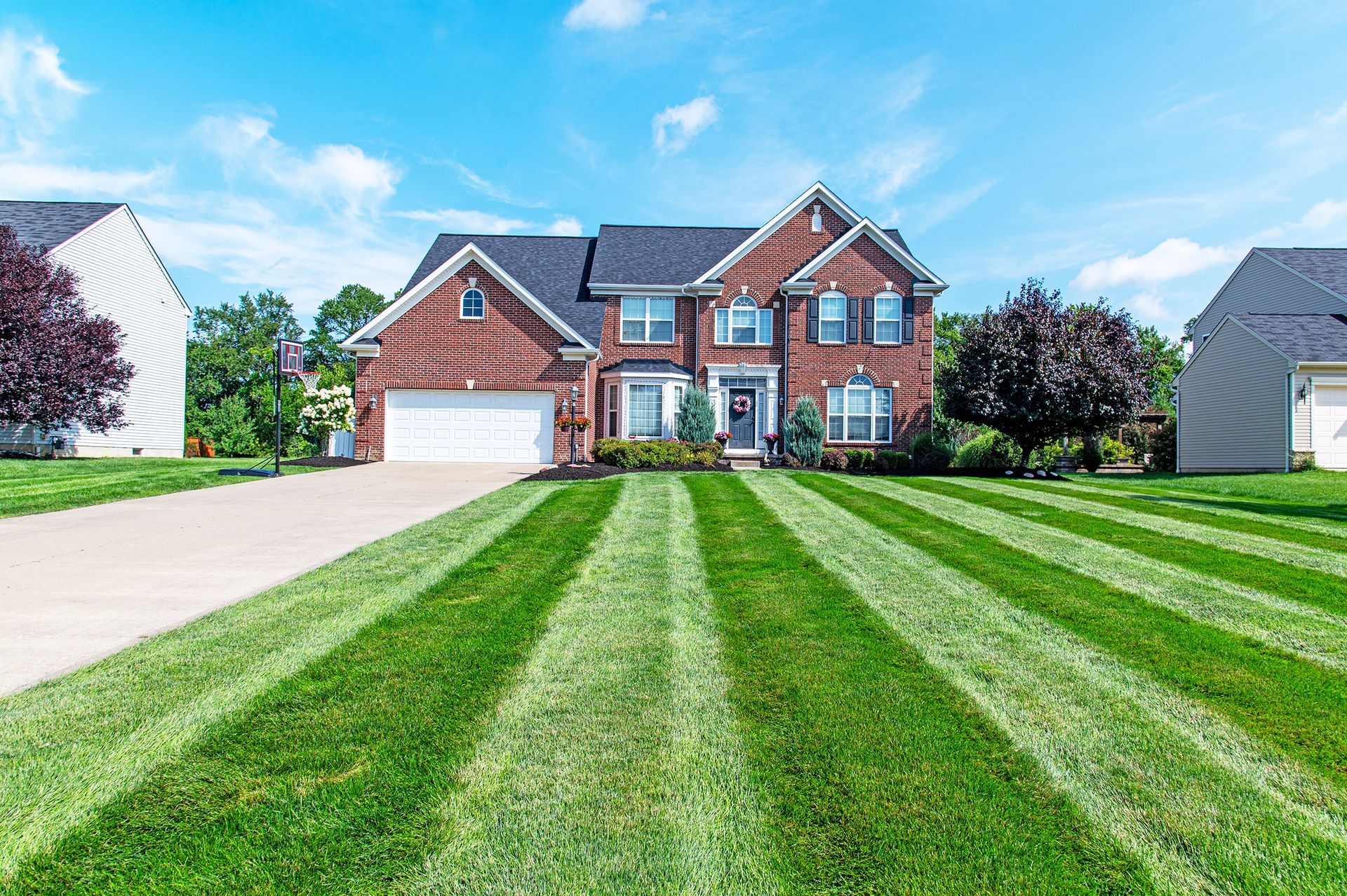 A two-story red brick suburban house with a white garage and a lawn featuring pronounced, symmetrical mowing stripes.