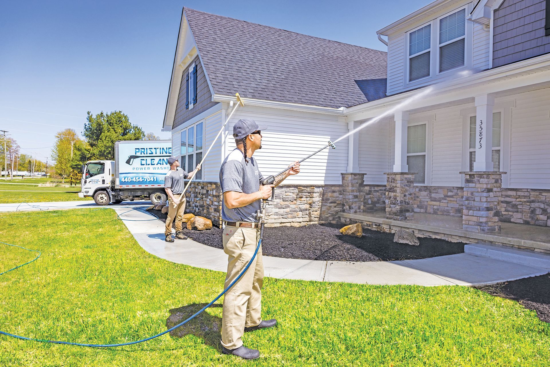 Two workers use extended pressure washing poles to clean the siding and soffits of a house on a sunny day.