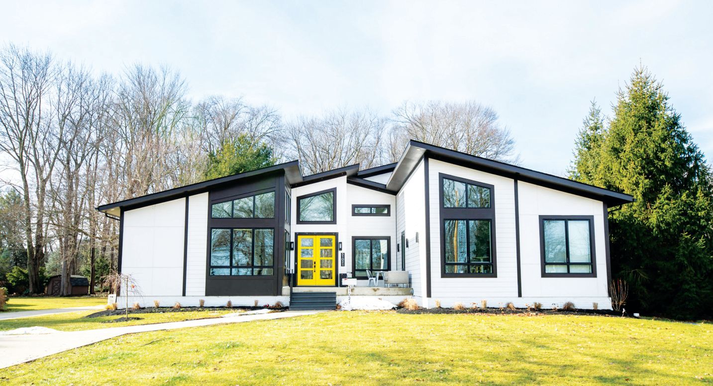 A modern, white single-story house with dark trim, large windows, and a vibrant yellow front door, set against a forest.