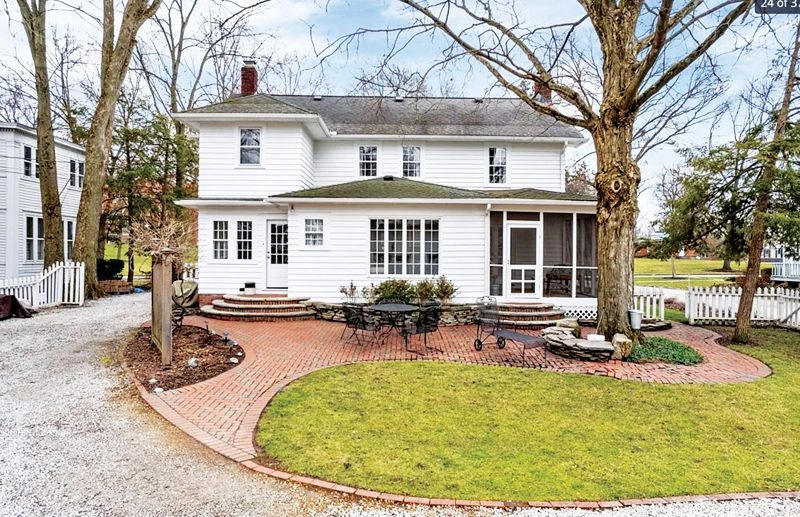 A two-story white house with a brick patio, a small lawn, and a gravel driveway on a bright, cloudy day.