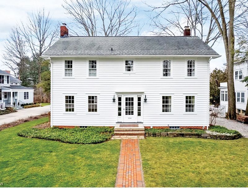 A two-story, white clapboard Colonial house with a central door and symmetrical windows, set behind a brick walkway.