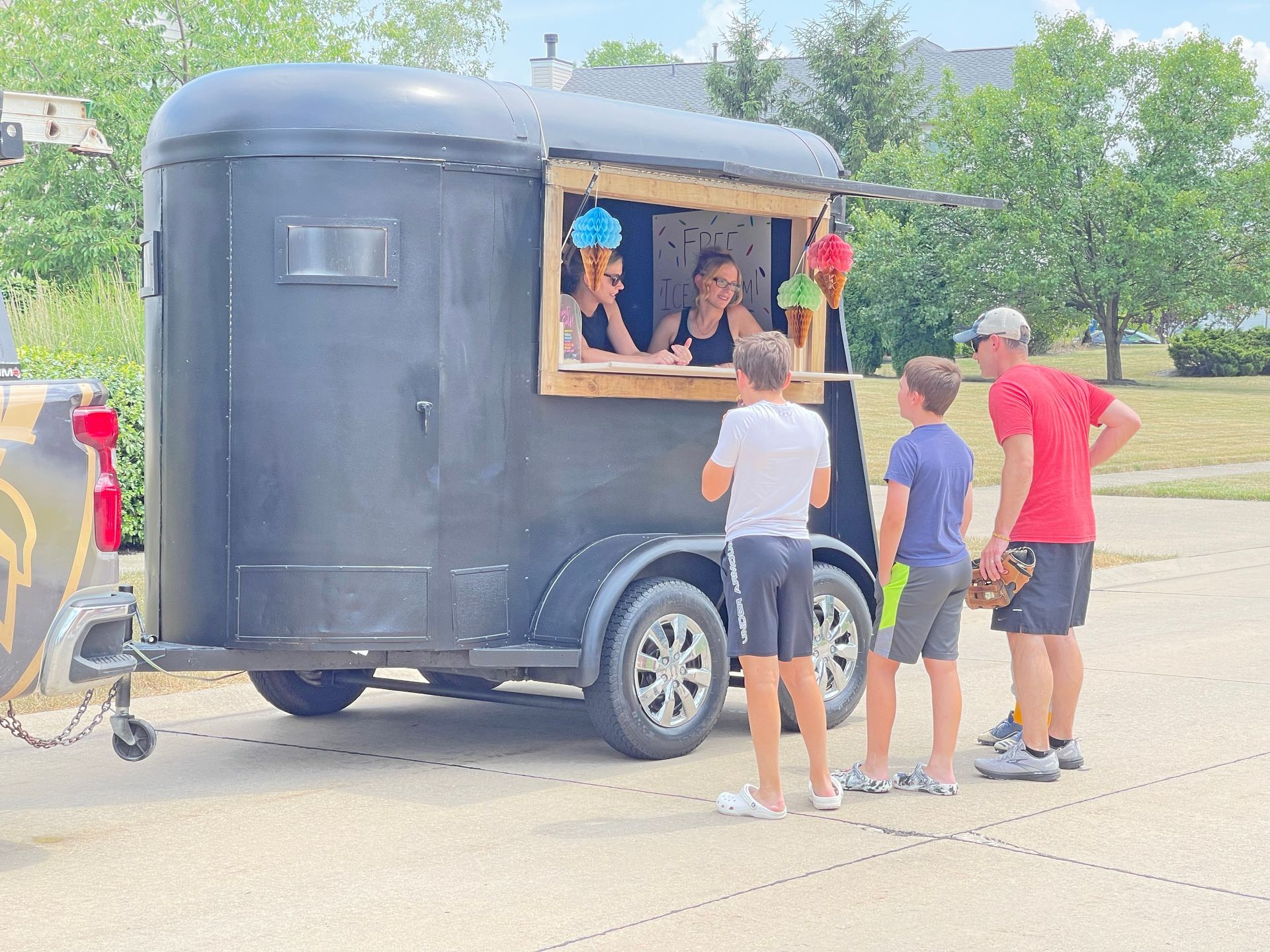 Two people wait at a mobile food stand inside a repurposed black horse trailer, while an adult stands to the side.