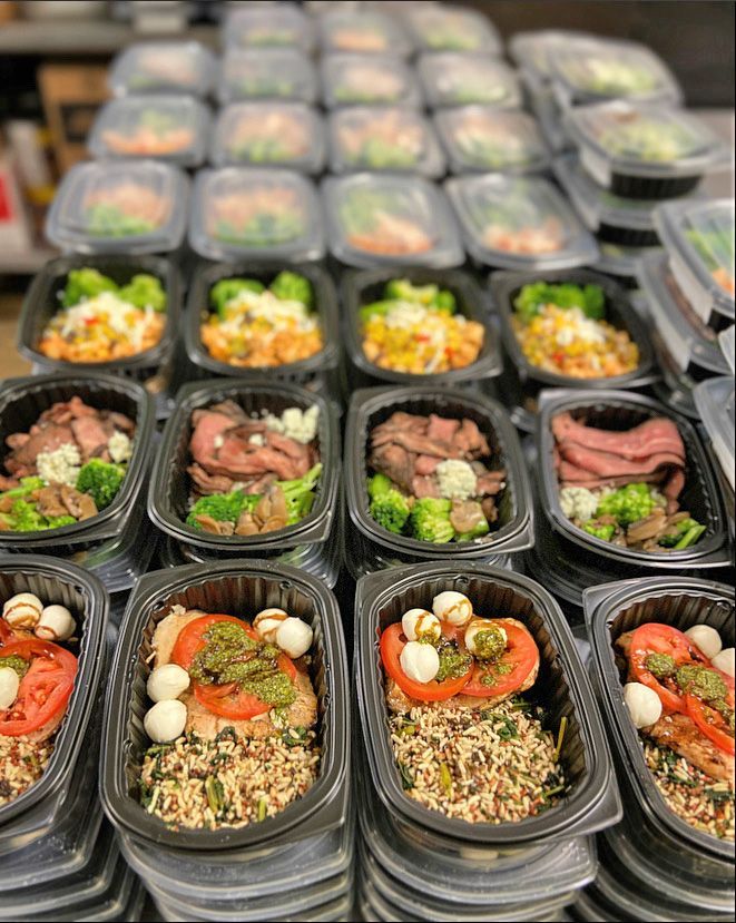 Rows of meal-prep containers filled with sliced meat, vegetables, rice, tomatoes, and mozzarella balls on a table.