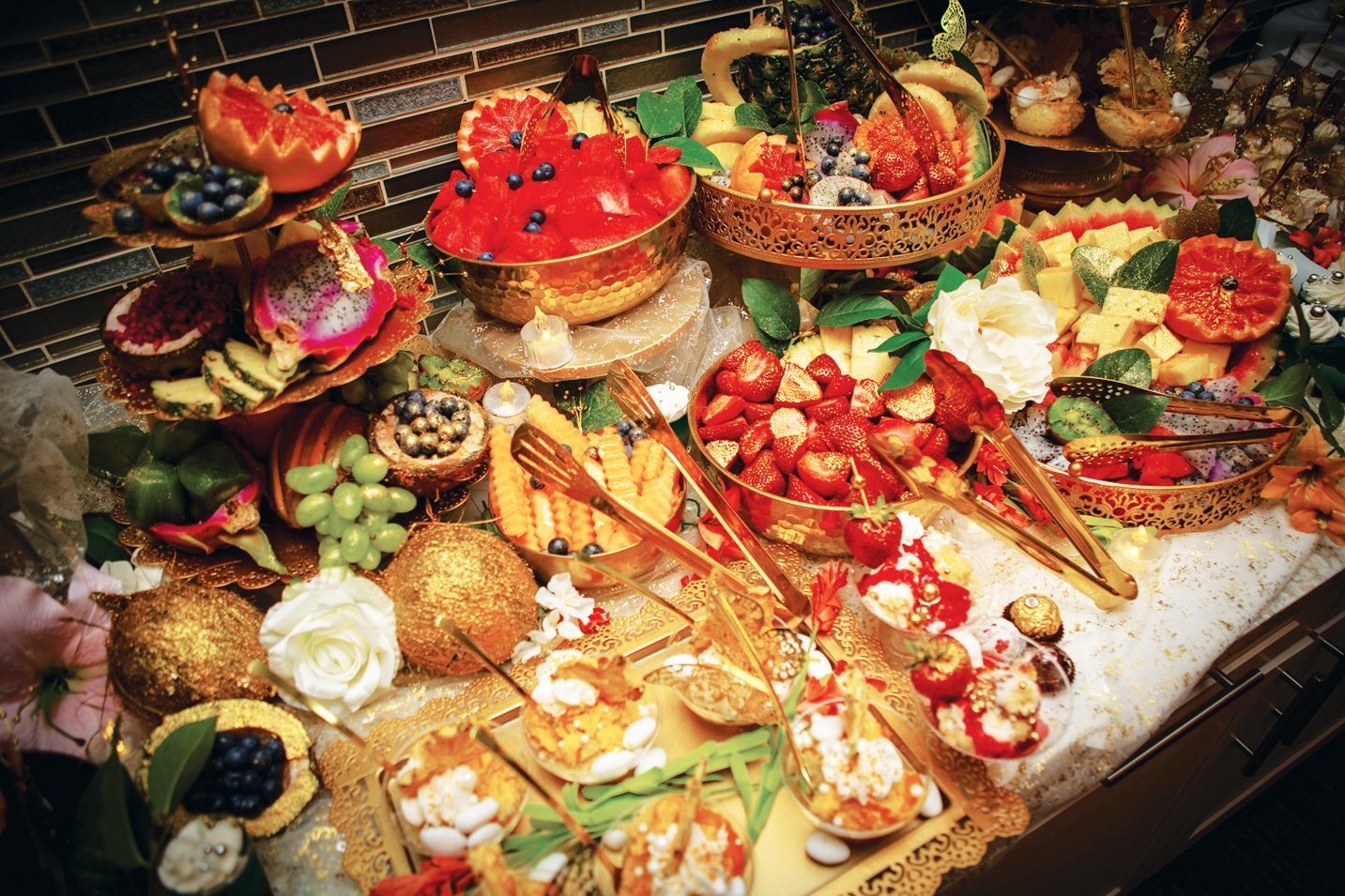 An elaborate fruit and hors d'oeuvre spread with bowls of strawberries, grapes, and citrus on a gold-accented table.