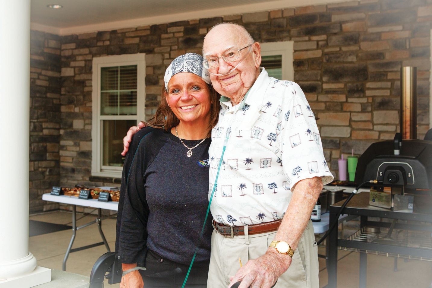 A smiling woman wearing a patterned head covering stands beside an elderly man in a patterned shirt on a stone patio.