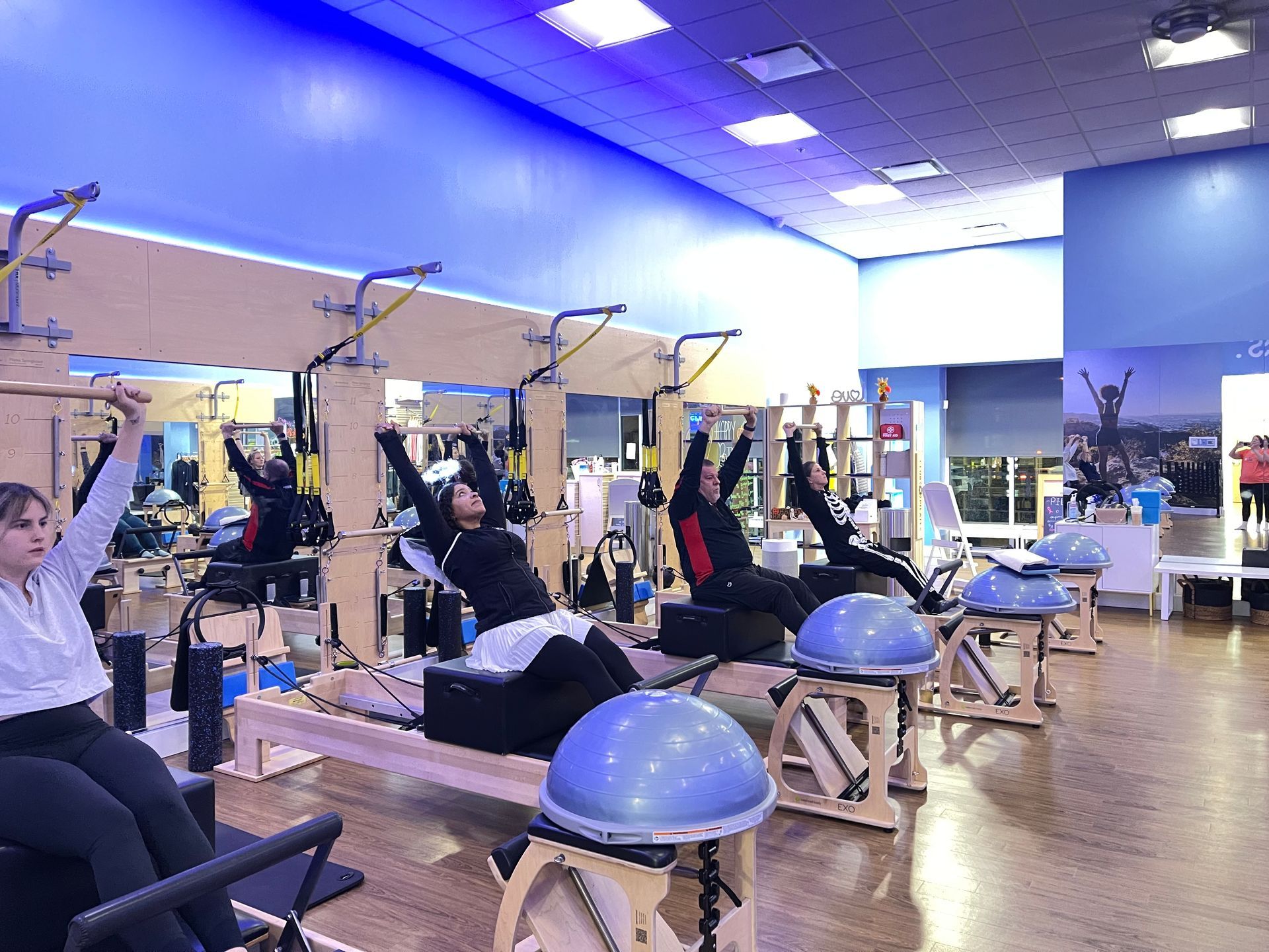 People in a studio exercising on Pilates reformer machines with blue BOSU balance balls in the foreground.