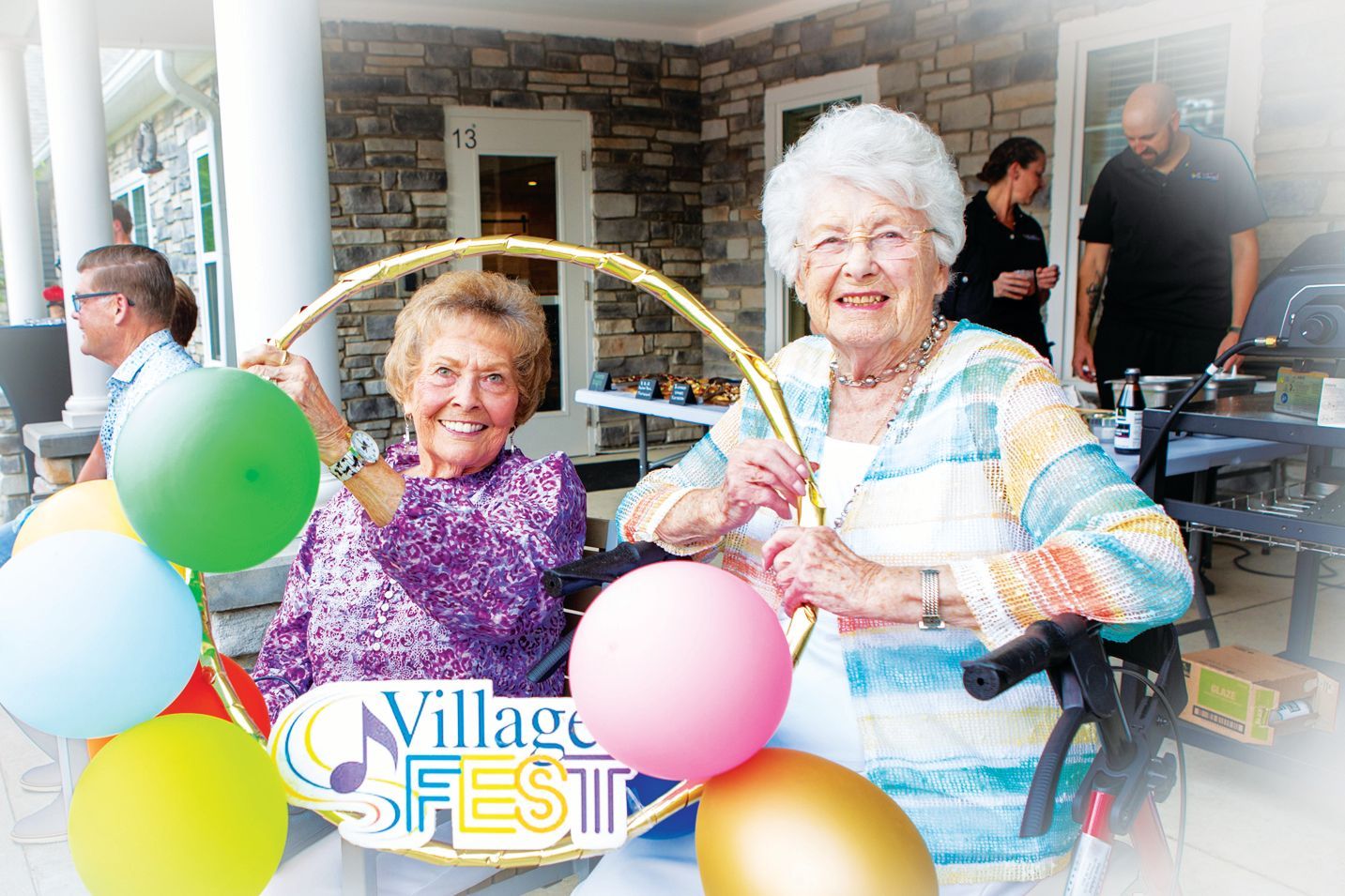 Two smiling people hold a gold hula hoop decorated with colorful balloons at an outdoor Village Fest event.