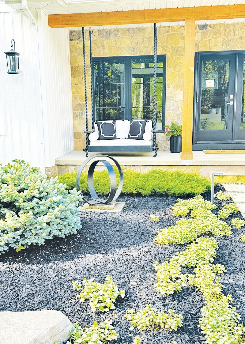 A modern home porch features a black swing with white cushions, situated behind a landscaped garden with dark mulch.