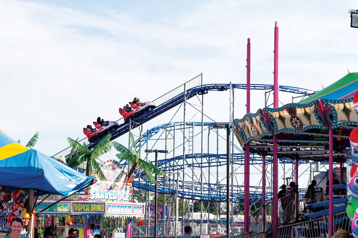 A blue roller coaster climbs a hill at a brightly colored carnival with a carousel and game booths in the foreground.
