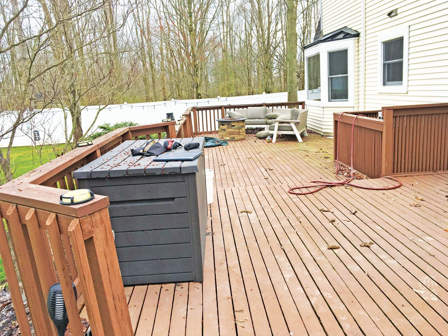 A wooden deck attached to a house with a black storage chest in the foreground and outdoor furniture in the background.