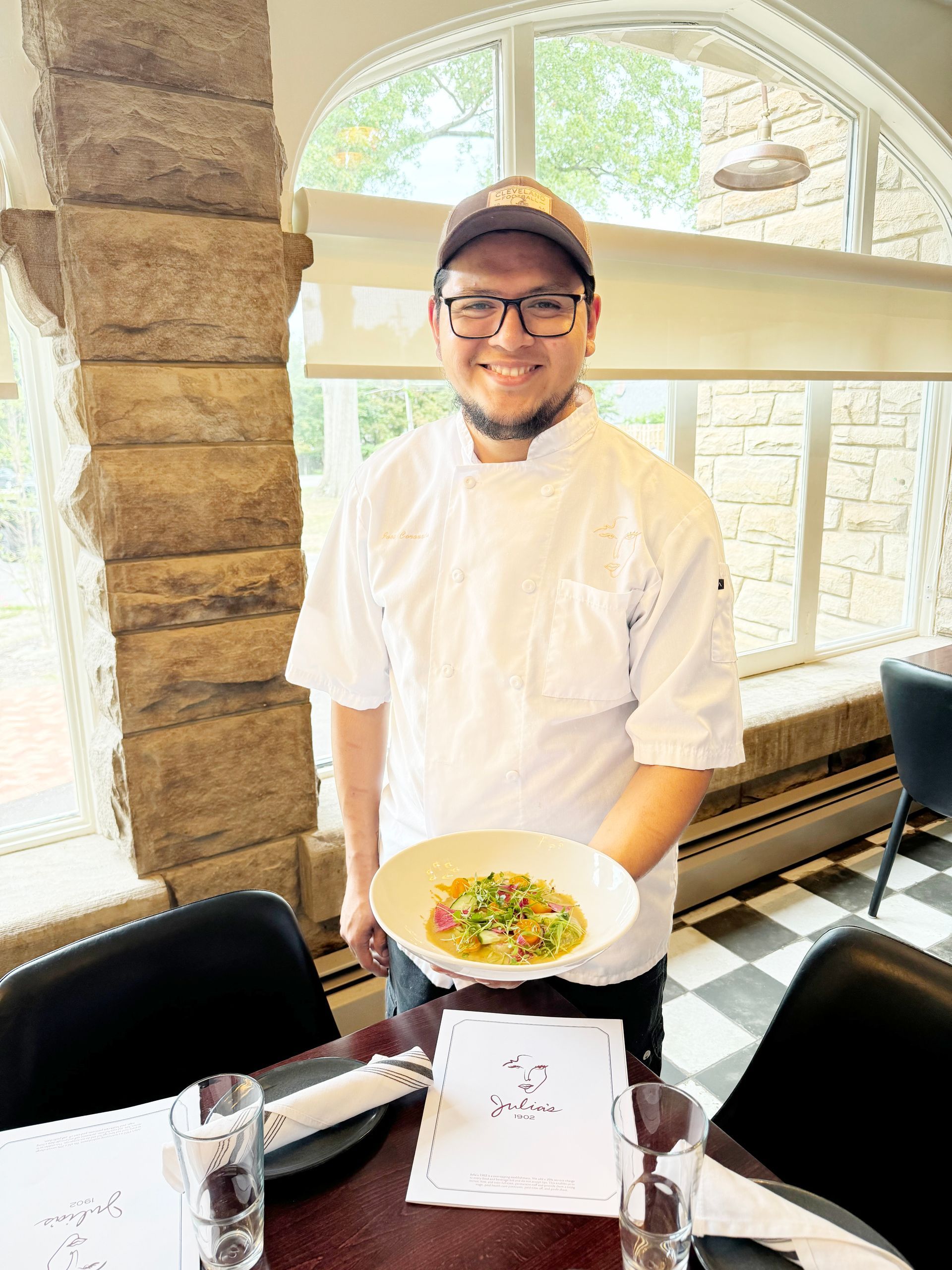 A chef in a white uniform holding a bowl of food in a restaurant setting with large windows and checkered flooring.
