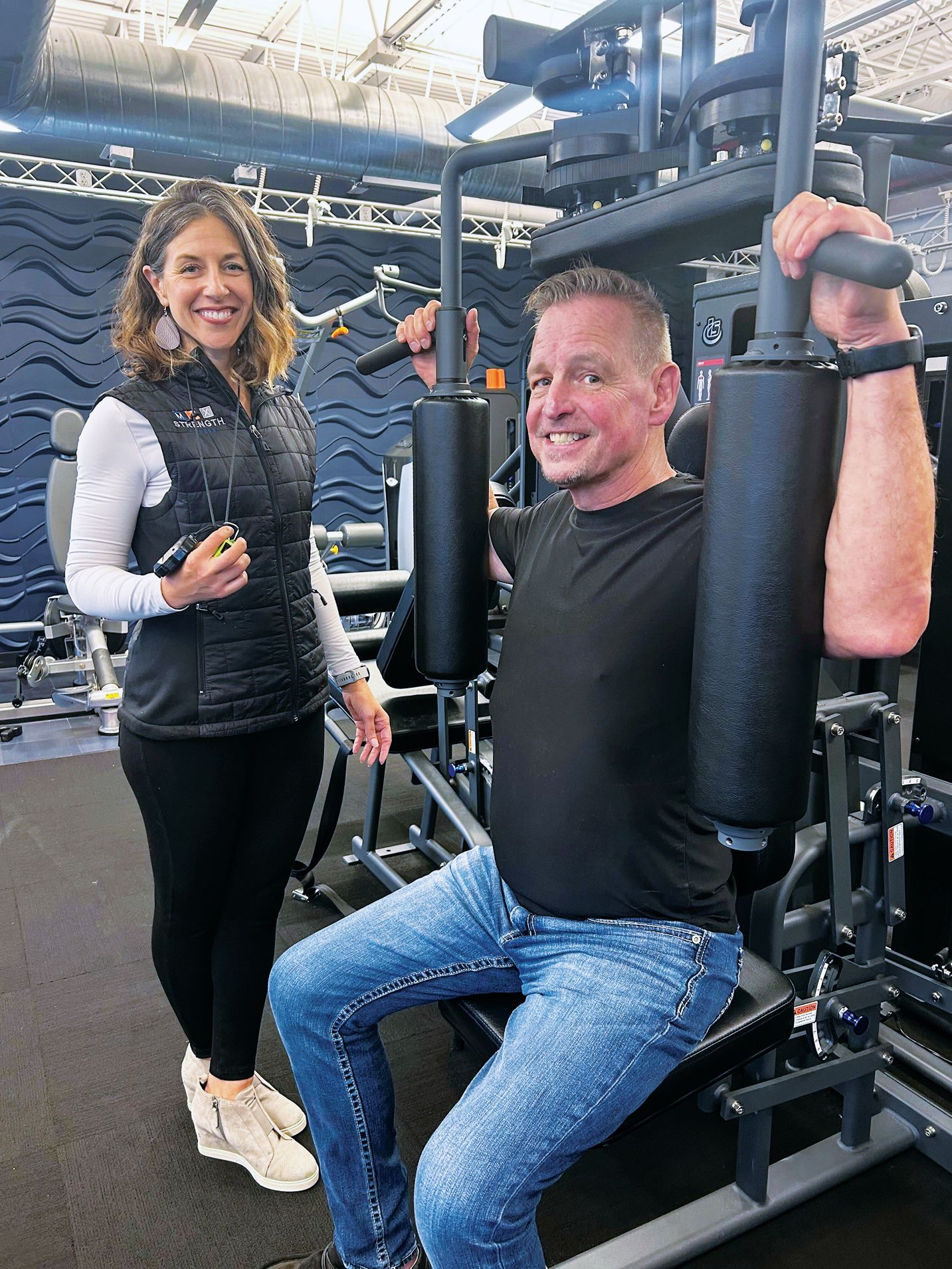 A smiling person sits at a gym machine as another person stands nearby, both inside a fitness facility.