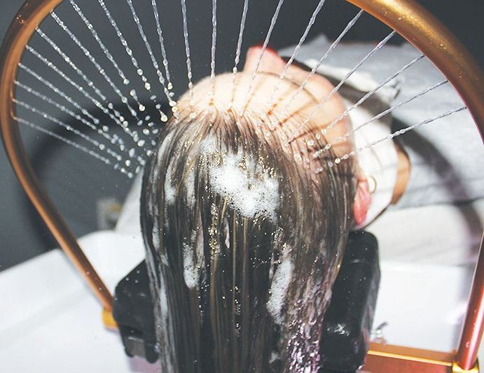 A person reclining in a salon sink as a curved, metallic shower fixture sprays water over their shampoo-covered hair.