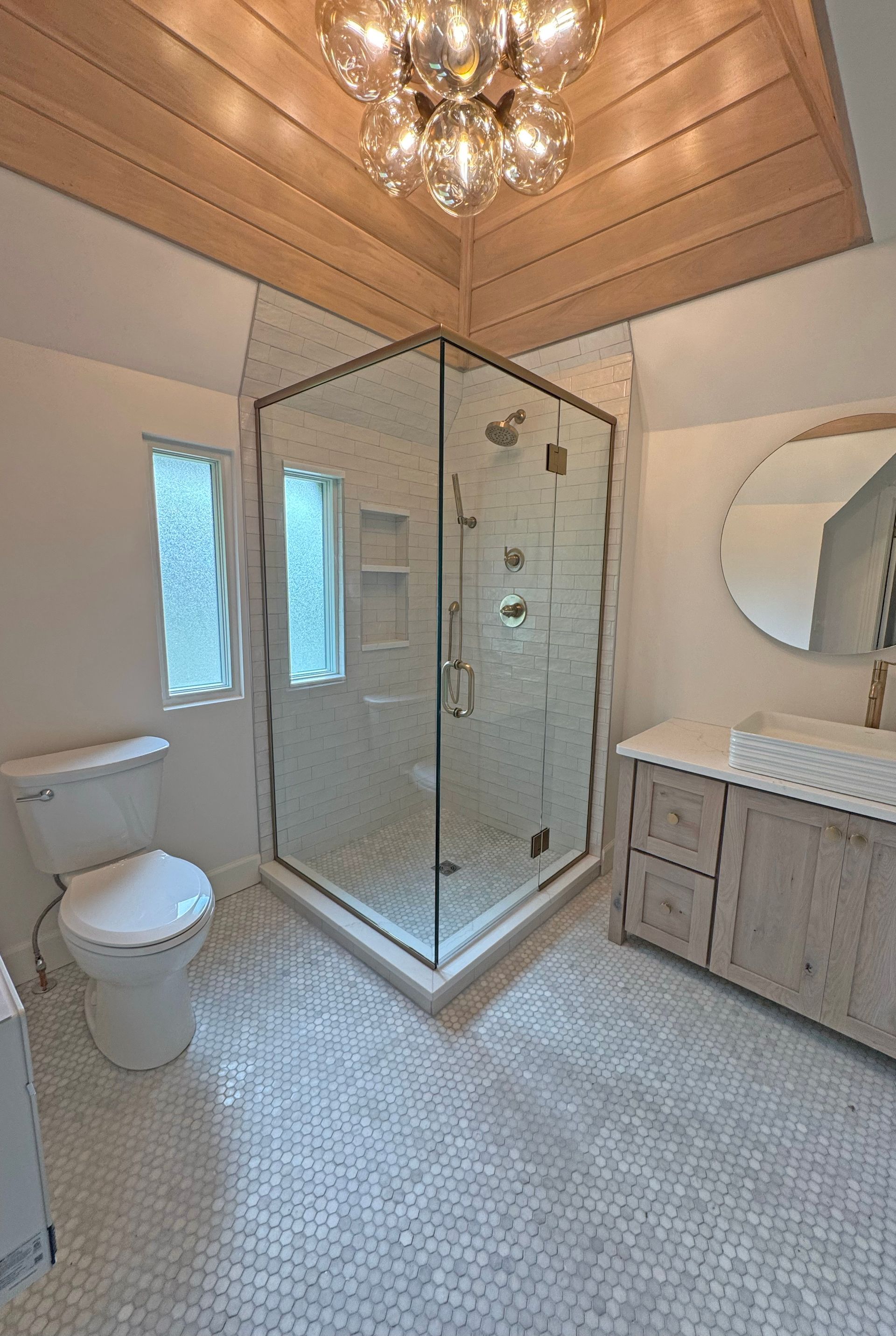 A modern bathroom with a glass corner shower, a white toilet, a light wood vanity, and a wooden paneled ceiling.