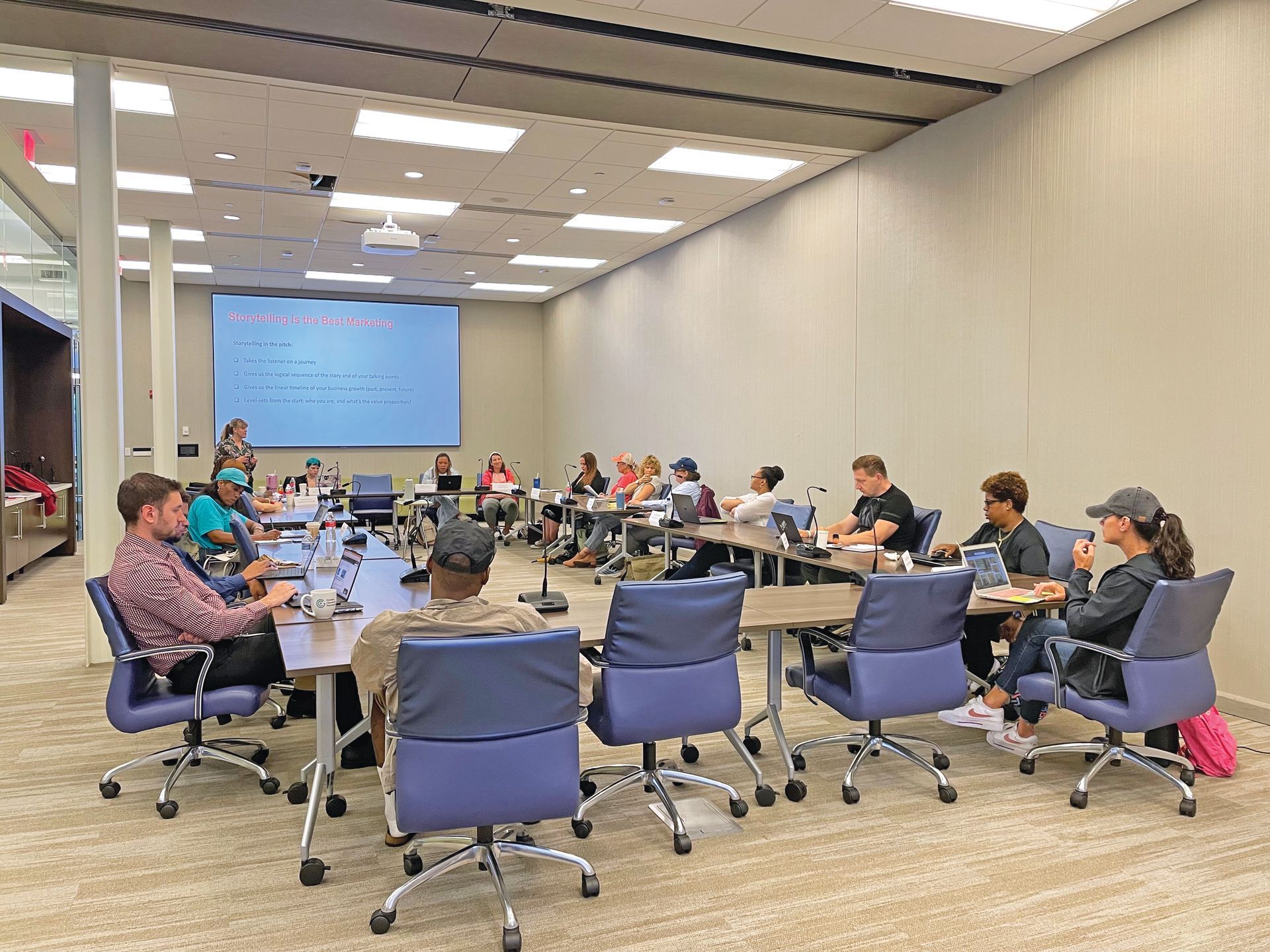 A group of people sit around a large U-shaped table in a bright, modern conference room, facing a screen for a meeting.