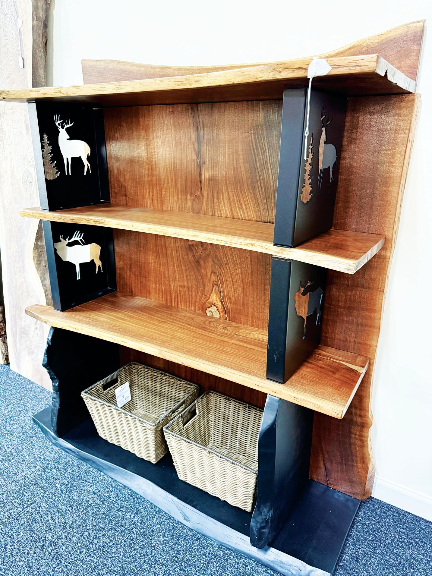 A three-tier, live-edge wood bookshelf with black vertical supports decorated with white deer silhouettes and two baskets.