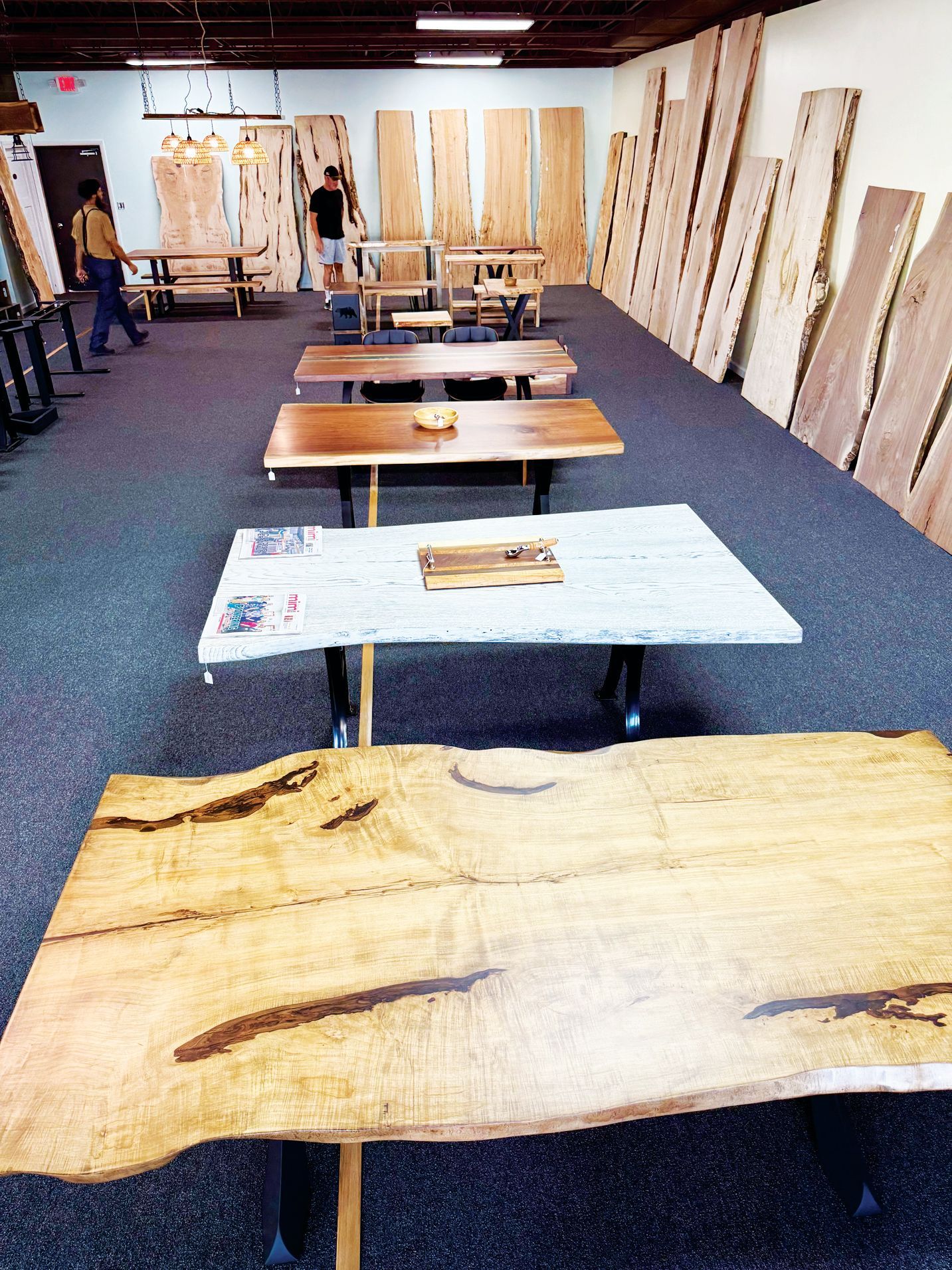 A showroom displays several live-edge wood slab tables arranged in a row, with wooden planks leaning against the wall.