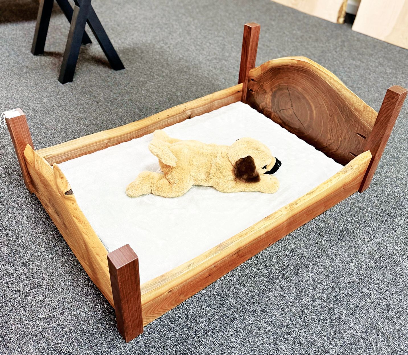A small, handcrafted wooden pet bed with a live-edge headboard, featuring a tan plush dog toy resting on a white cushion.