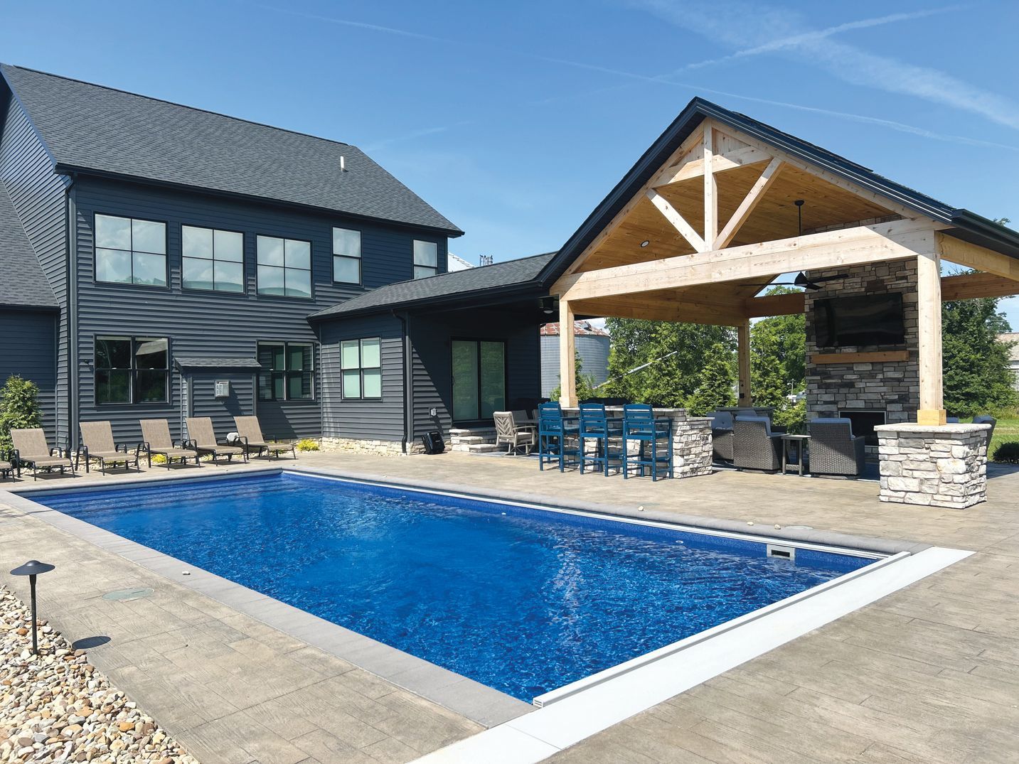 A rectangular backyard swimming pool beside a modern black house with an adjacent open-air wood pavilion and patio area.