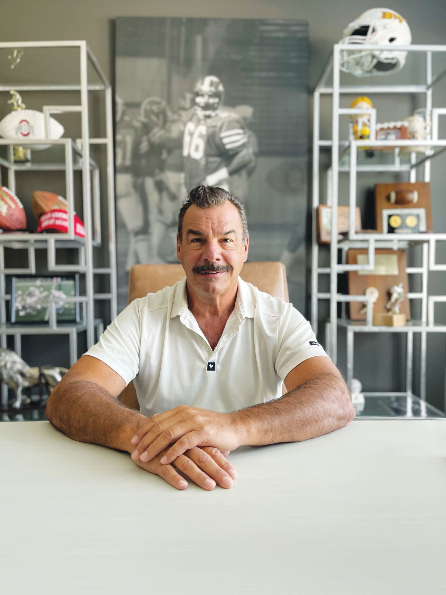 A man with a mustache sits at a desk with his hands clasped, in front of a sports-themed office with shelves and memorabilia.