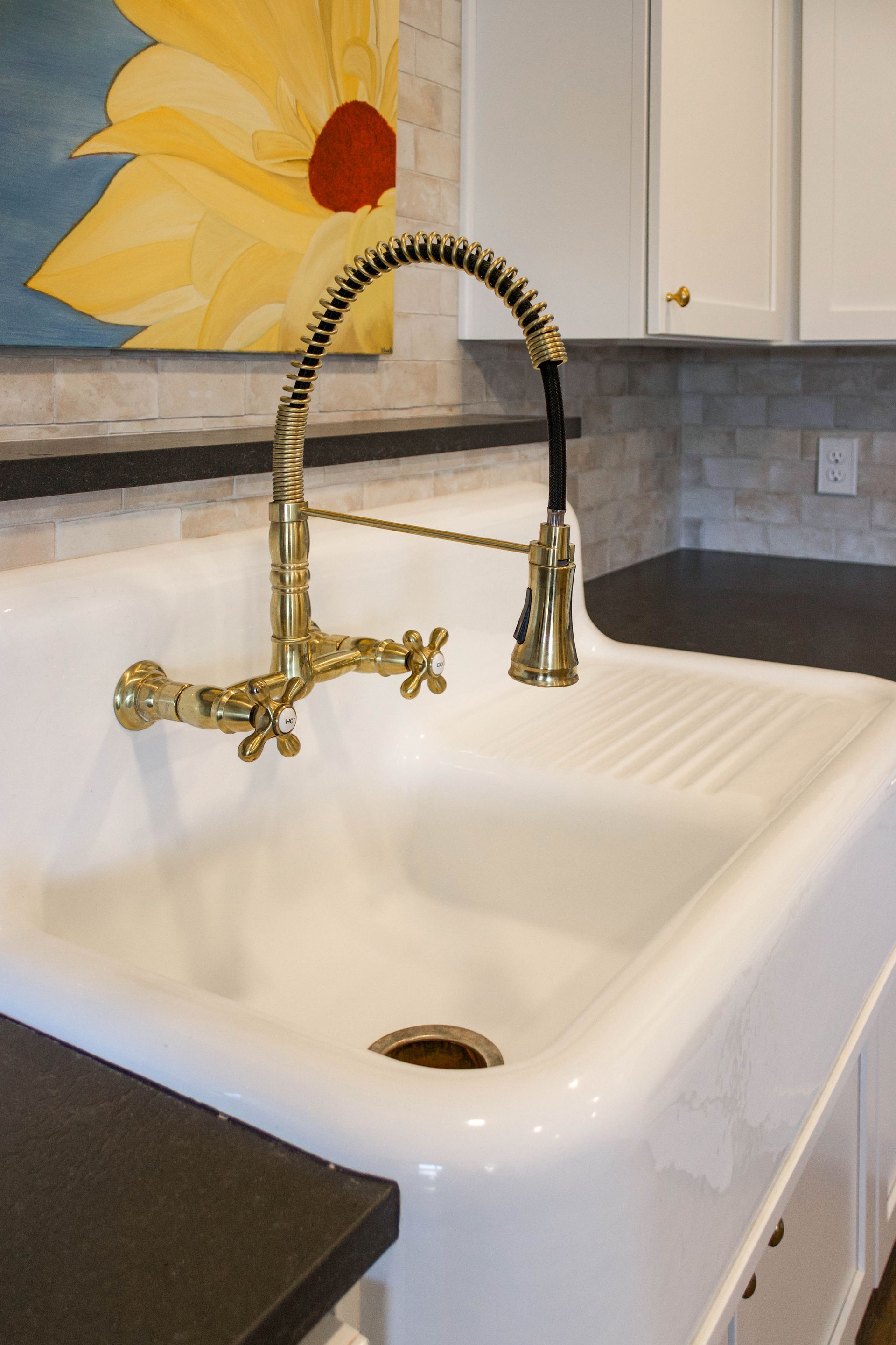 A brass, industrial-style kitchen faucet mounted on a large, white farmhouse sink with a drainboard.