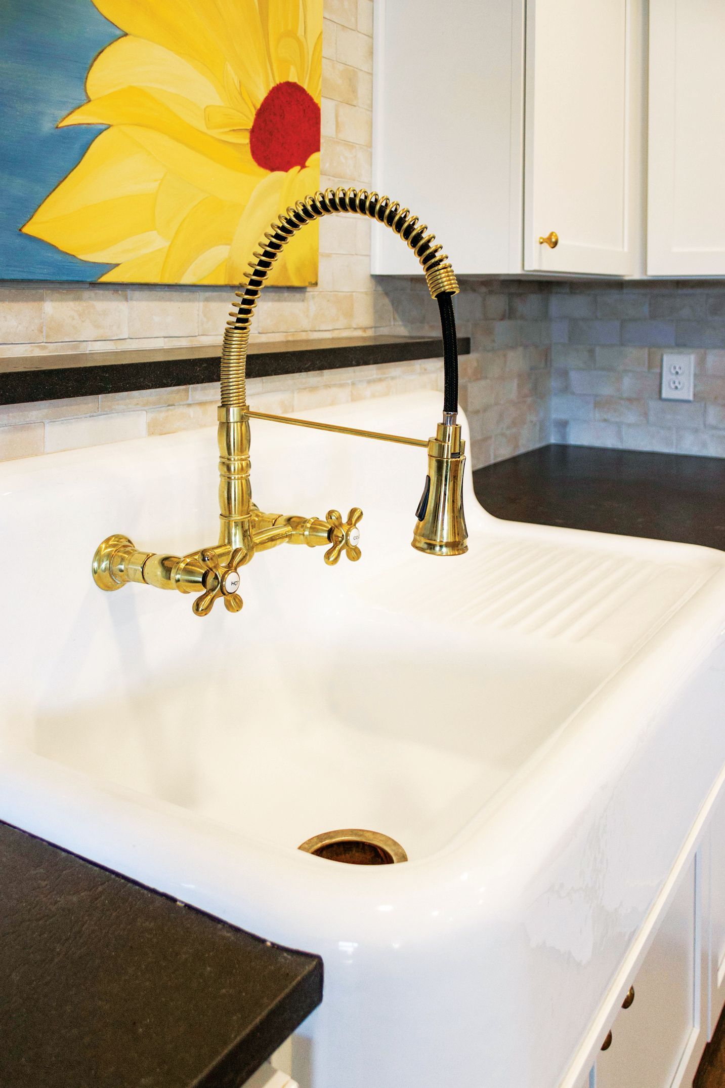 A white farmhouse sink with a gold gooseneck faucet, set against a stone backsplash and a yellow sunflower painting.
